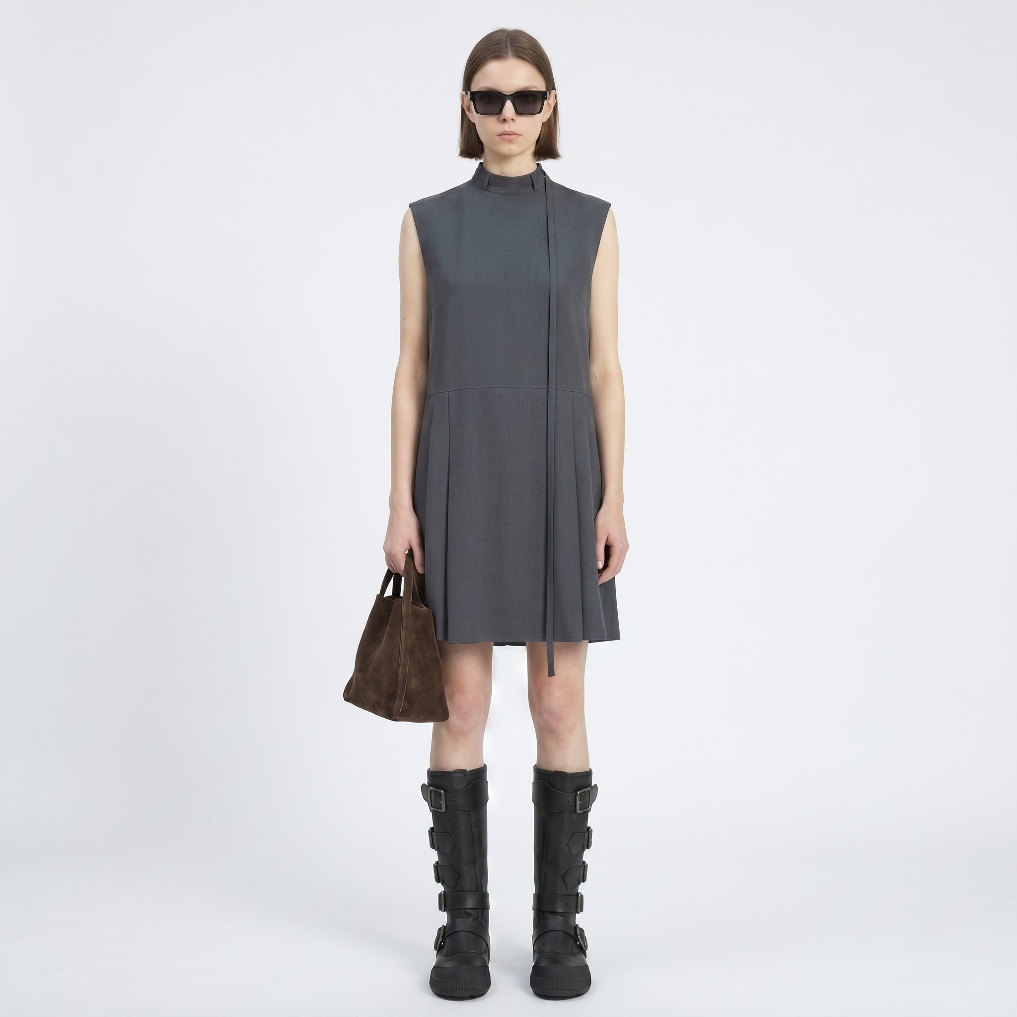 Woman wearing a sleeveless gray dress with black boots and sunglasses on a white background